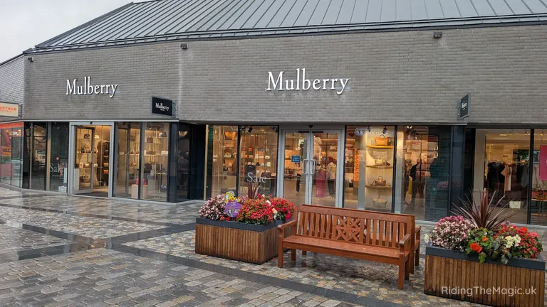 Mulberry shopfront with glass display windows, flower planters and a wooden bench in a paved shopping area.