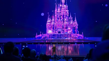 Illuminated fairytale castle projected in pink and purple, with a performer on stage before an audience.