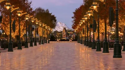 Illuminated pedestrian avenue lined with autumn trees and ornate street lamps, leading towards snow-capped mountains at dusk.