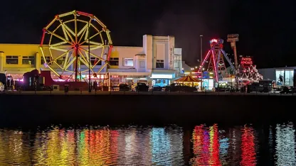 Night-time amusement rides, including a Ferris wheel, beside a waterfront with colourful reflections.