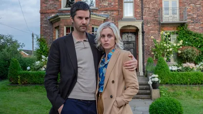 A man and woman stand together outside a brick house, with the man’s arm around the woman.