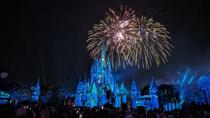 Fireworks illuminate a blue-lit fairytale castle at night, with a crowd watching in the foreground.