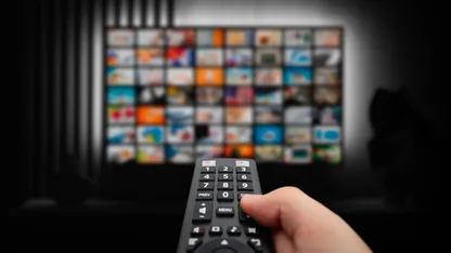 Hand holding a remote control aimed at a television displaying a grid of channels.