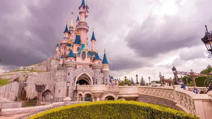 Disney-style fairytale castle with pink turrets and blue roofs beneath dark, cloudy skies.