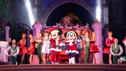 Mickey Mouse and Miinnie Mouse in Father Christmas style outfits arms open with dancers dressed in red, white and green coloured clothes on stage with arms open wide