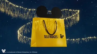 Yellow shopping bag with black handle and black Mickey Mouse ears coming out of the top. Writing in botoom of thirs of yellow bag is black: Selfridges with black mickey mouse emoji Blue starlight background with zig zag of gold glitter shooting through background