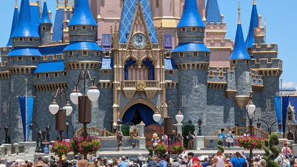 grey castle with blue tiled turrets and pink building behind lamp posts with two orb white lights hanging baskets pink flowers tops of peoples heads in crowd