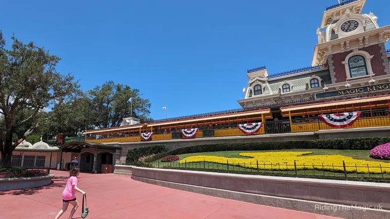 Magic Kingdom railway station with a clock tower, bunting and landscaped gardens.