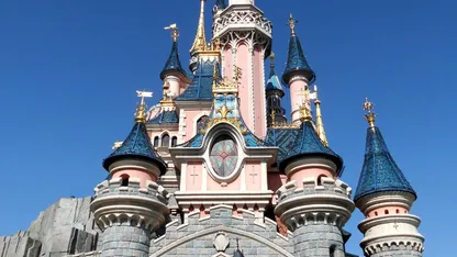 Pink fairytale castle with blue turrets and ornate stone towers against a clear blue sky.