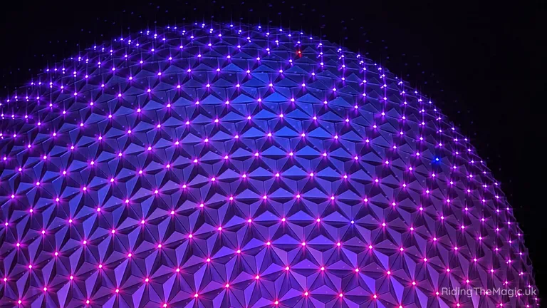 Purple-lit geodesic dome with a repeating triangular metal façade and glowing lights against a dark background.