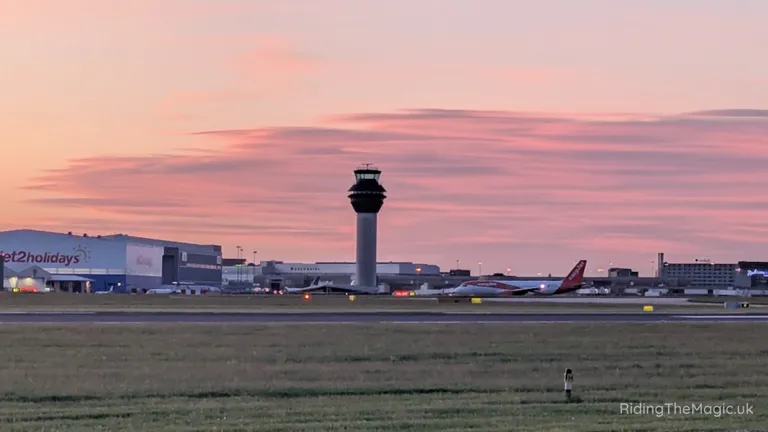 Airport control tower and aeroplane on the tarmac at sunset, viewed across grassy grounds.