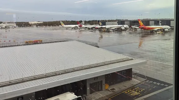 Airport apron with several parked aircraft on a wet tarmac, viewed from a terminal window.