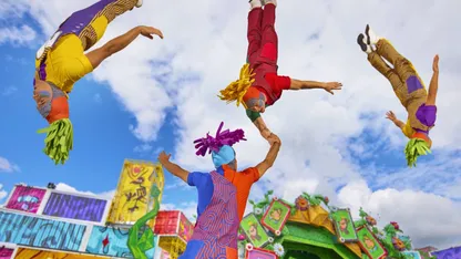 Four brightly costumed acrobats perform outdoors, with three suspended upside down around a central performer.