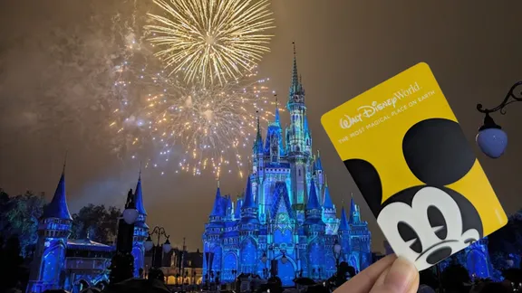 Fireworks explode over the blue‑lit Cinderella Castle at Walt Disney World as a hand holds a Mickey-themed park card in the foreground.