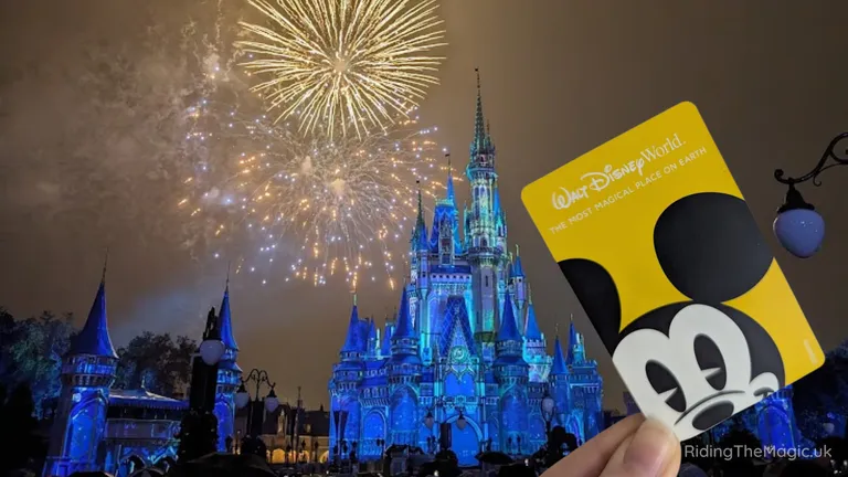 Fireworks explode over the blue‑lit Cinderella Castle at Walt Disney World as a hand holds a Mickey-themed park card in the foreground.