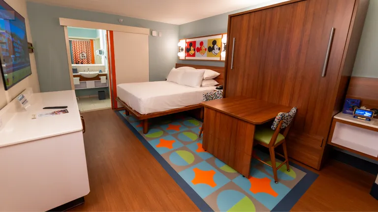 Hotel room with a double bed, wooden wardrobe, desk and colourful patterned rug, decorated with Mickey Mouse artwork.