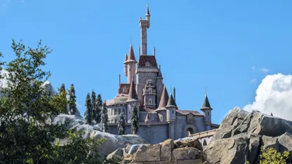 Fairytale castle with multiple towers and spires, surrounded by trees and rocky landscape under a blue sky.
