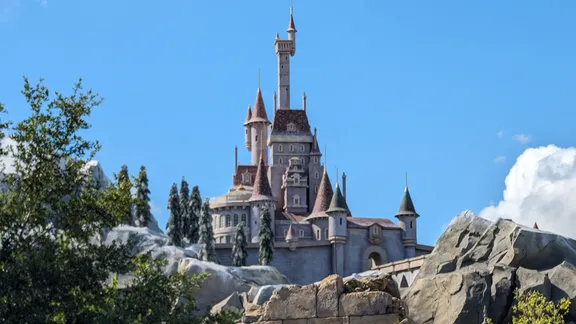 Fairytale castle with multiple towers and spires, surrounded by trees and rocky landscape under a blue sky.