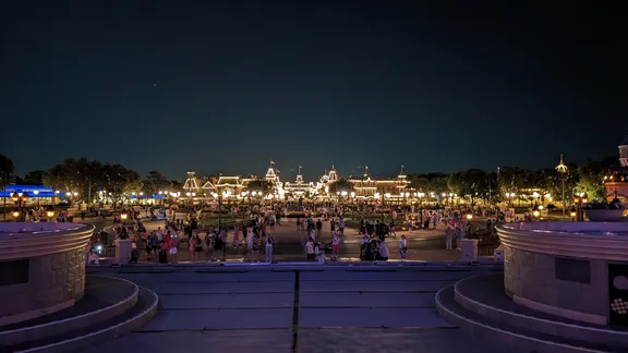 Night-time view of Main Street, U.S.A., at a lit theme park, with crowds gathered in the central plaza.