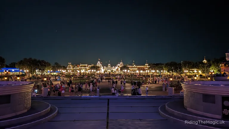 Night-time view of Main Street, U.S.A., at a lit theme park, with crowds gathered in the central plaza.