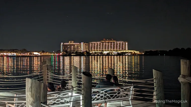 Lit waterfront hotel at night, reflected in the water behind a dock with people seated on benches.