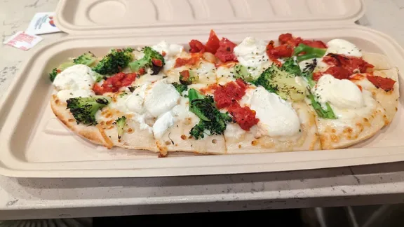 Flatbread pizza topped with mozzarella, tomatoes, broccoli and herbs in a takeaway container.