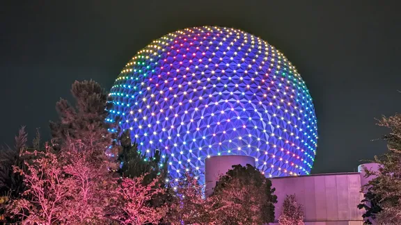 Illuminated EPCOT geodesic sphere glowing with blue, purple and green lights at night, surrounded by trees.