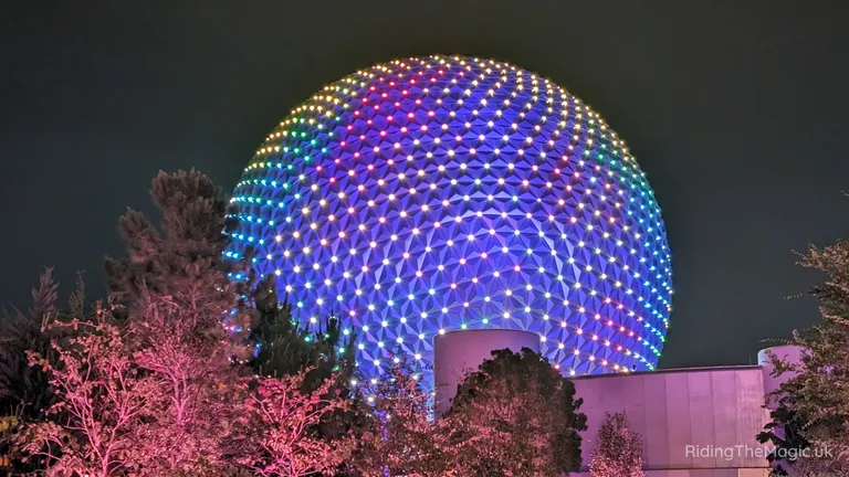 Illuminated EPCOT geodesic sphere glowing with blue, purple and green lights at night, surrounded by trees.