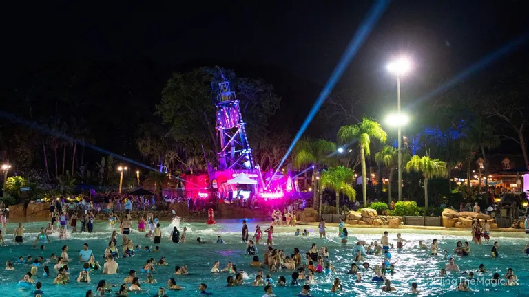 Crowded outdoor swimming pool at night, illuminated by colourful lights and beams around a central stage.