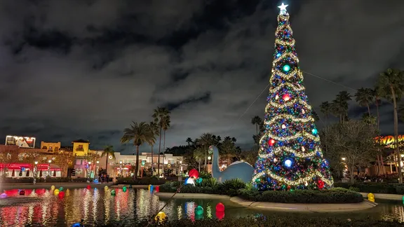 Night-time view of a brightly decorated Christmas tree beside a reflective pond, with palm trees and shops in the background.