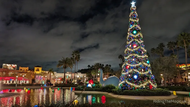 Night-time view of a brightly decorated Christmas tree beside a reflective pond, with palm trees and shops in the background.