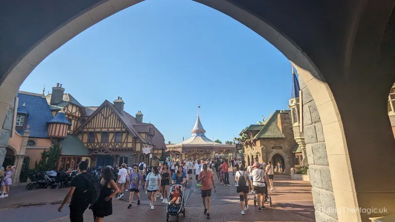 Crowded theme park street with Tudor-style buildings and a carousel, viewed through a stone archway.