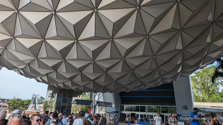 Crowd gathered beneath the large geometric, faceted canopy at a Spaceship Earth building EPCOT entrance.