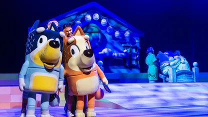 Two costumed animal characters stand on a blue-lit stage, with performers and a house-themed set behind them.