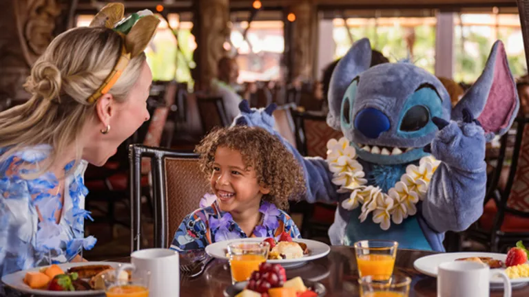 Child smiles at breakfast with a woman and Stitch character wearing a lei at a restaurant table.