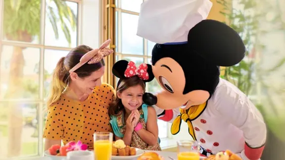 A girl celebrates with a chef-costumed Mickey Mouse character and an adult at a breakfast table.