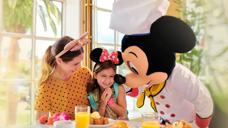 A girl celebrates with a chef-costumed Mickey Mouse character and an adult at a breakfast table.