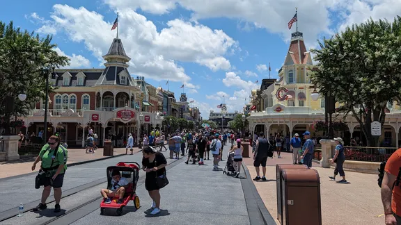 Crowded theme park entrance Main Street USA with ornate Victorian-style buildings, American flags, and visitors walking beneath a blue sky.