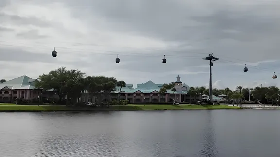 Cable cars crossing above a lake, with pastel-coloured buildings and trees along the far shore under a cloudy sky.