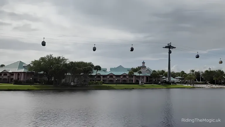 Cable cars crossing above a lake, with pastel-coloured buildings and trees along the far shore under a cloudy sky.