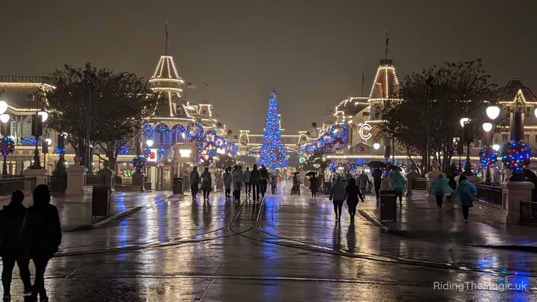 People walking down a wet, holiday-lit street toward a large blue Christmas tree at night.