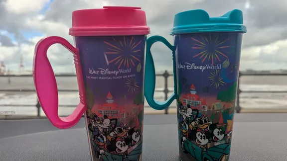 Two Disneyland World travel mugs, one pink and one teal, on a table by the water.