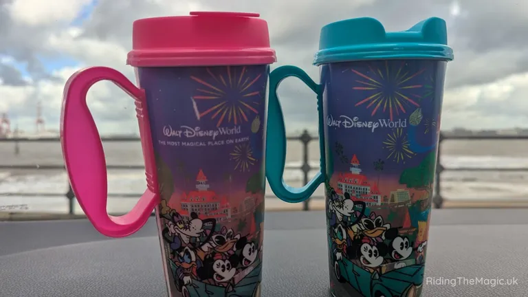 Two Disneyland World travel mugs, one pink and one teal, on a table by the water.