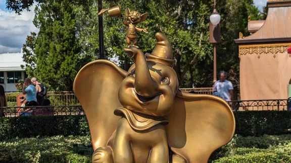 Golden Dumbo statue with a smiling expression, surrounded by trees, hedges and railings in an outdoor theme park.
