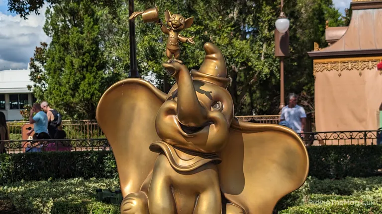 Golden Dumbo statue with a smiling expression, surrounded by trees, hedges and railings in an outdoor theme park.