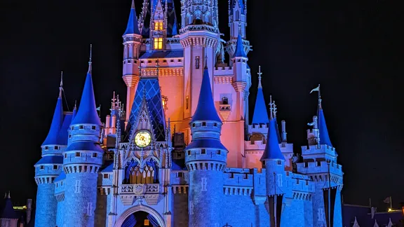 A brightly lit fairytale castle illuminated in blue and pink against a night sky.