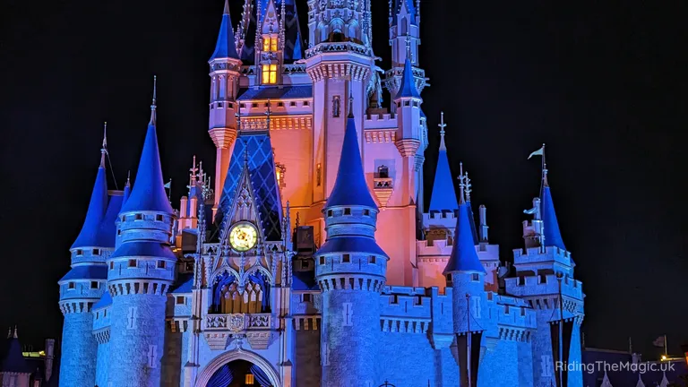 A brightly lit fairytale castle illuminated in blue and pink against a night sky.
