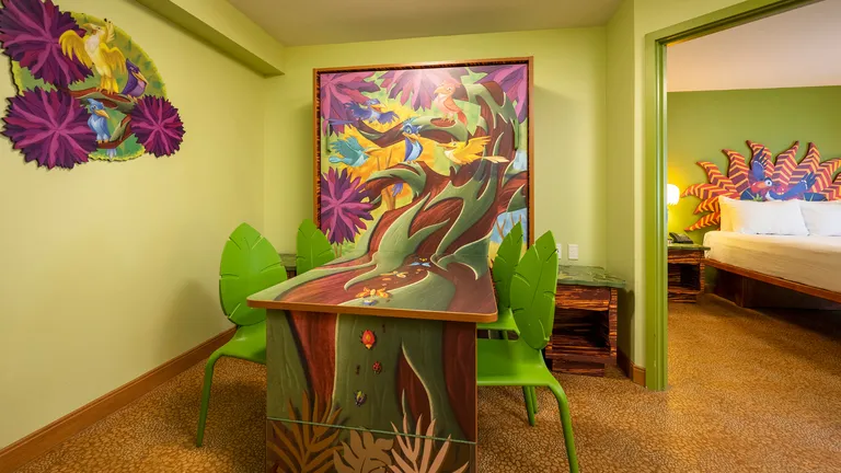 Tropical-themed room with a painted desk, two green chairs, colourful wall art and an adjoining bedroom.