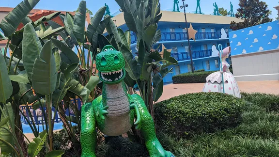 Green dinosaur statue in tropical landscaping outside a colourful hotel, with a rabbit sculpture and blue balconies in the background.