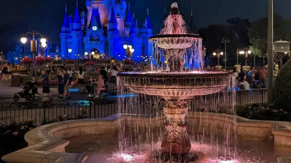 Cinderella Castle illuminated blue behind a tiered fountain at night in a crowded theme park.